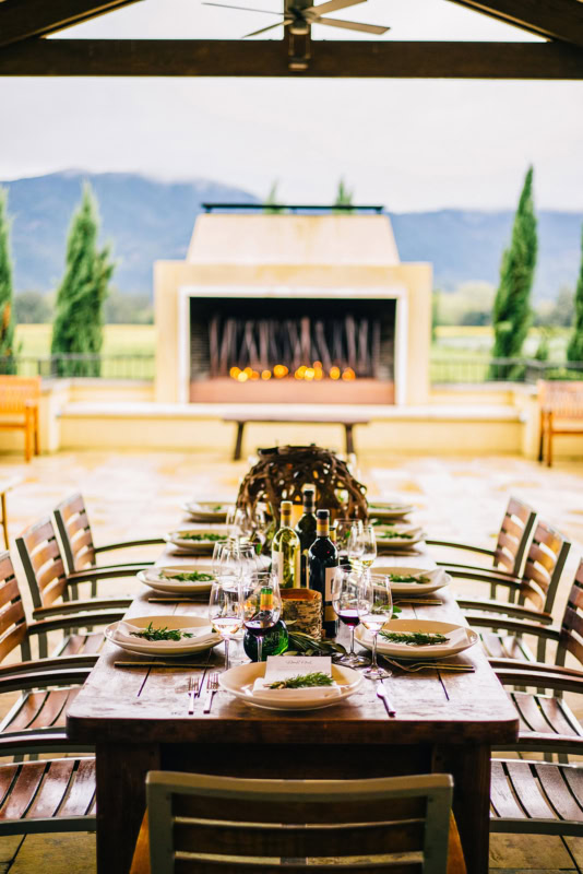 Elegant outdoor dining setup at Round Pond Estate with a long table, wine bottles, and a fireplace backdrop.
