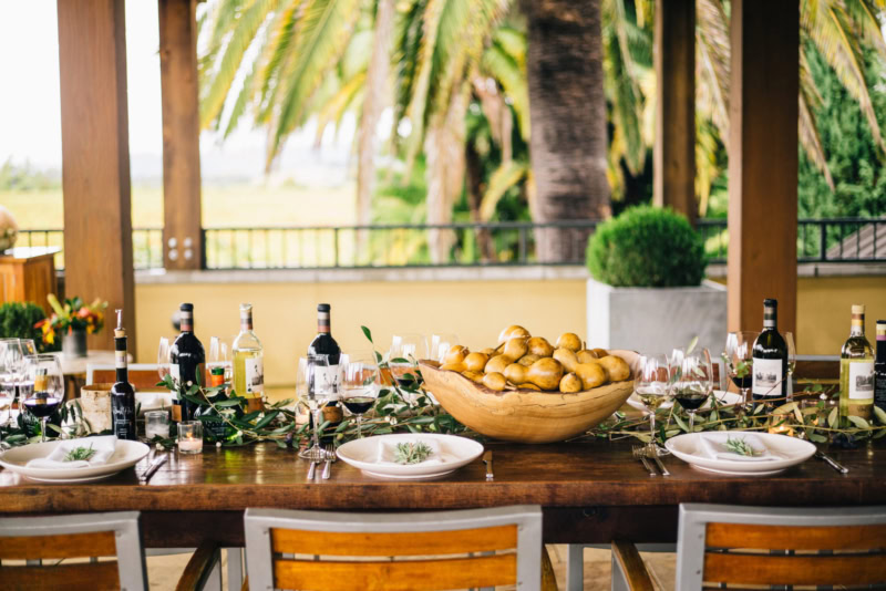 Elegant outdoor dining table at Round Pond Estate with wine bottles, glasses, and a large bowl of potatoes, surrounded by pal.