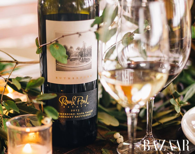 A bottle of red wine from Round Pond Estate with a glass of white wine in the foreground, surrounded by greenery and candles.