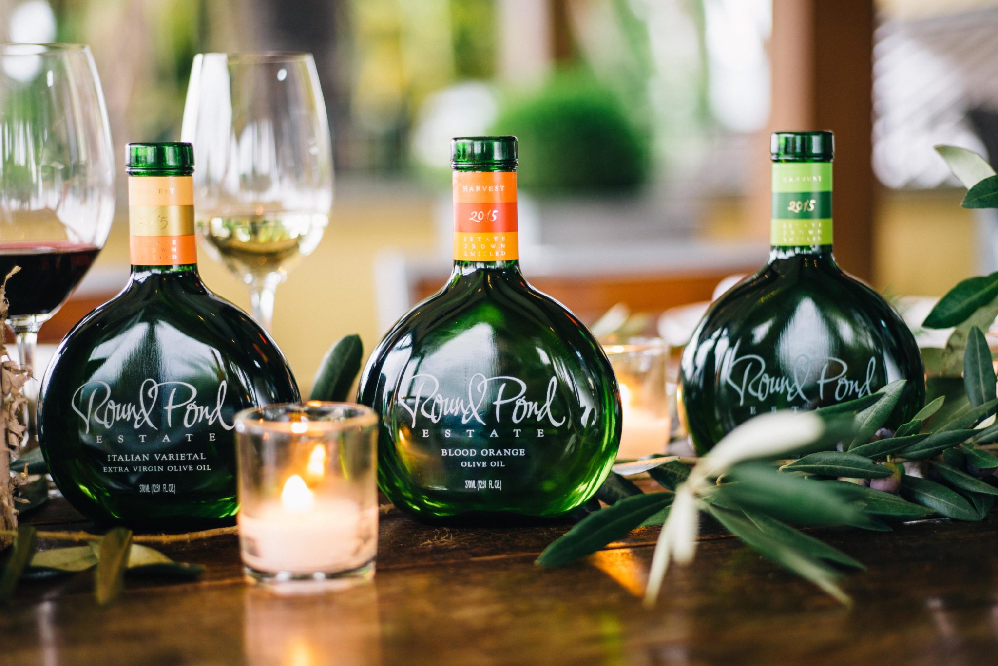 Three Round Pond Estate wine bottles with colorful labels, set on a wooden table with wine glasses and greenery in the backgr.