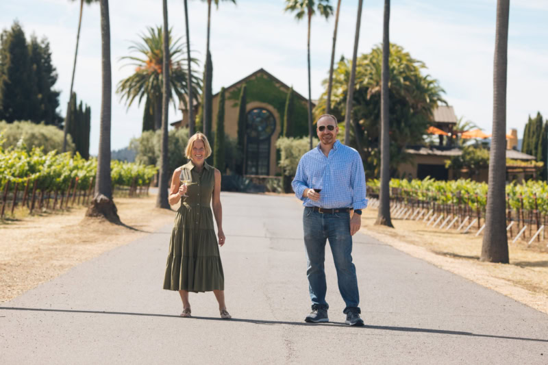 Two people standing on a vineyard path at Round Pond Estate with palm trees and a church in the background.