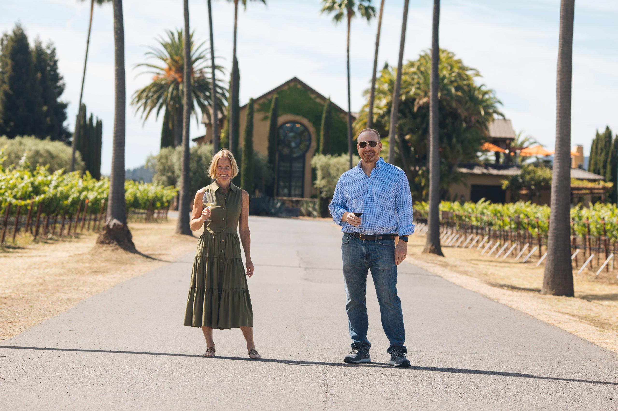Two people standing on a vineyard path at Round Pond Estate with palm trees and a church in the background.