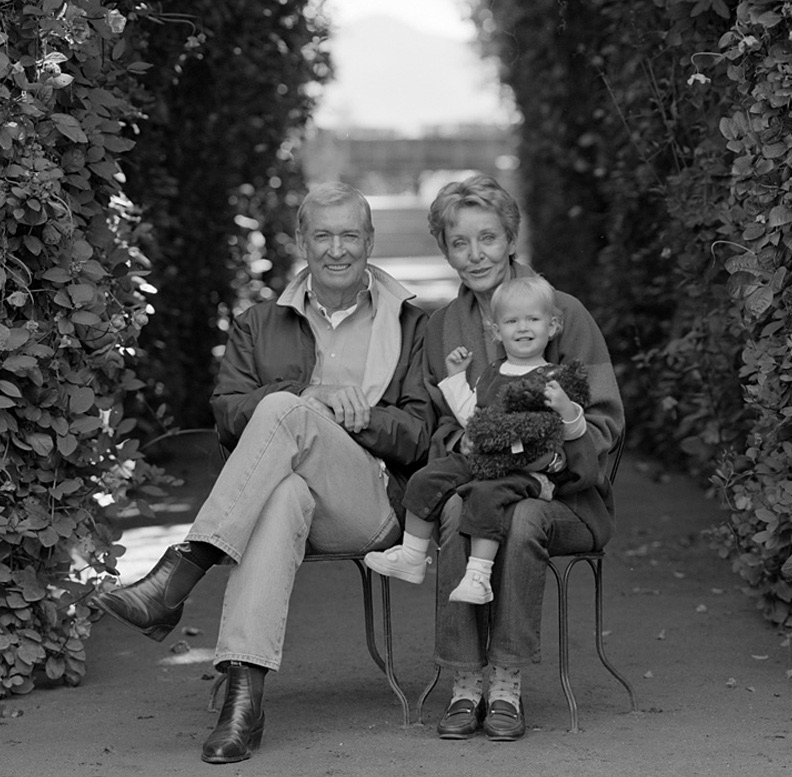 Family sitting on a bench in a garden pathway surrounded by lush bushes.