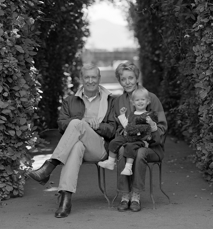 Black and white photo of three generations of a family sitting on chairs outdoors, surrounded by lush foliage.