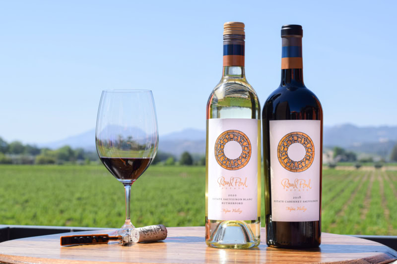 Two bottles of Napa Valley wine and a glass of red wine on a wooden table with vineyard background.