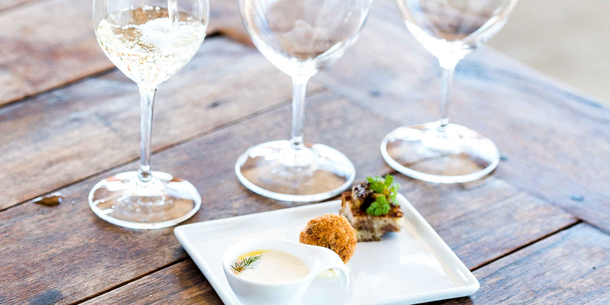 White wine being poured into a wine glass next to two empty wine glasses alongside a plate with three bite-sized pairings