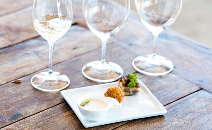 White wine being poured into a wine glass next to two empty wine glasses alongside a plate with three bite-sized pairings