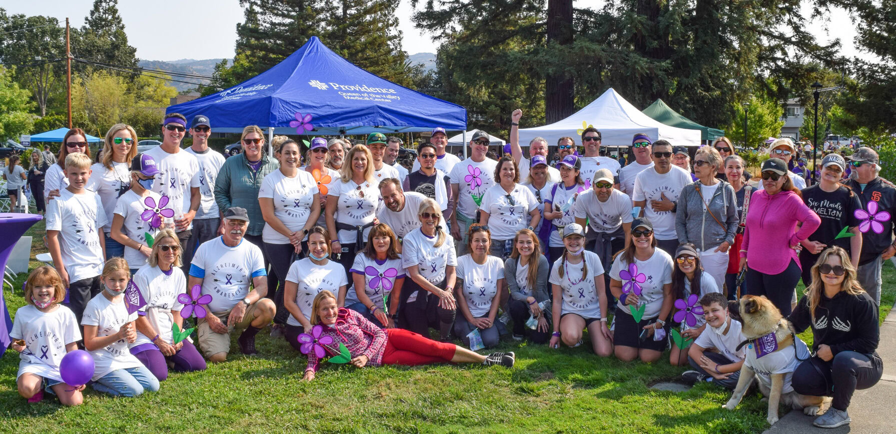 Group of diverse people, children, and dogs at an outdoor event with tents and trees in the background.