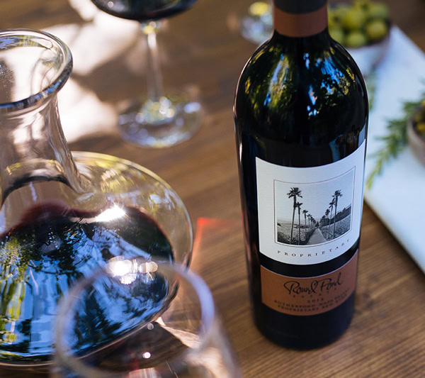 A bottle of red wine on a wooden table with glasses and a cheese platter, emphasizing wine and food pairing.