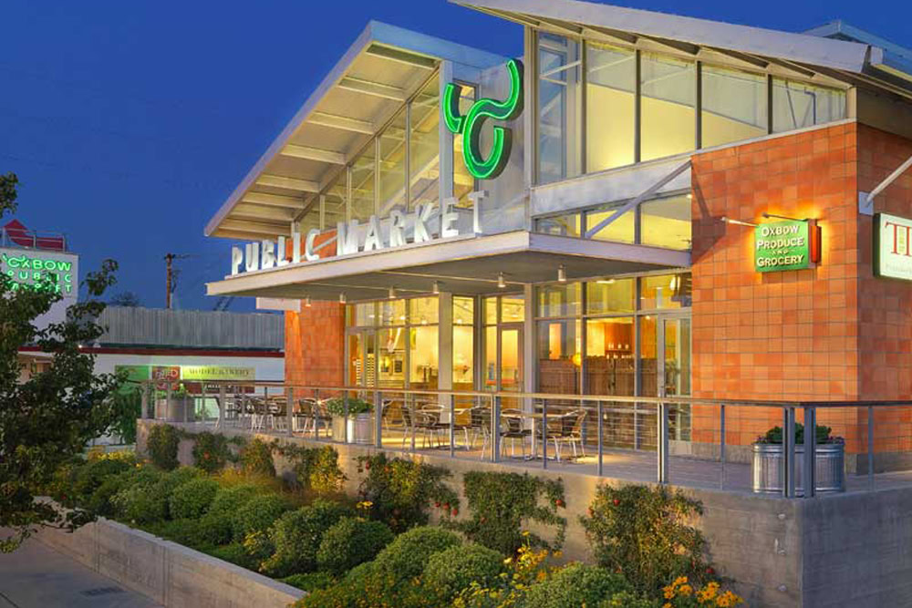 Brightly lit public market building at dusk with a green logo and large windows.