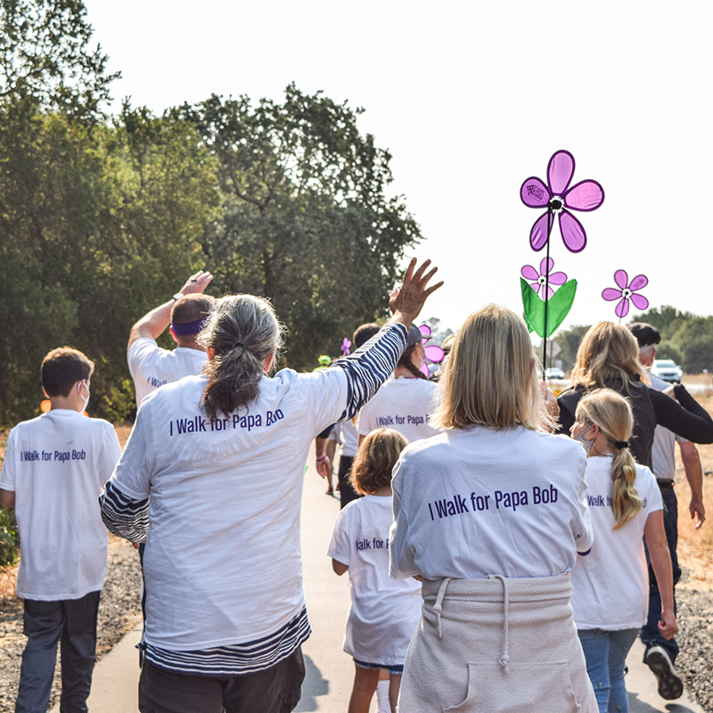 People walking to end Alzheimer's wearing white shirts with "I walk for Papa Bob" on the back and holding purple pinwheel flowers