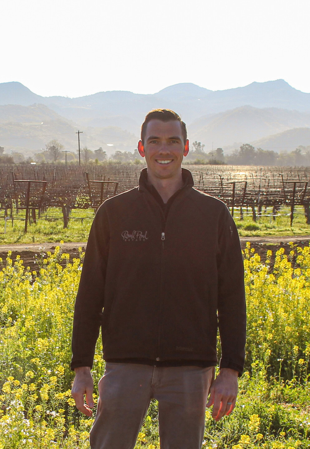 Man standing in a vineyard with mountains in the background, smiling, during daylight.