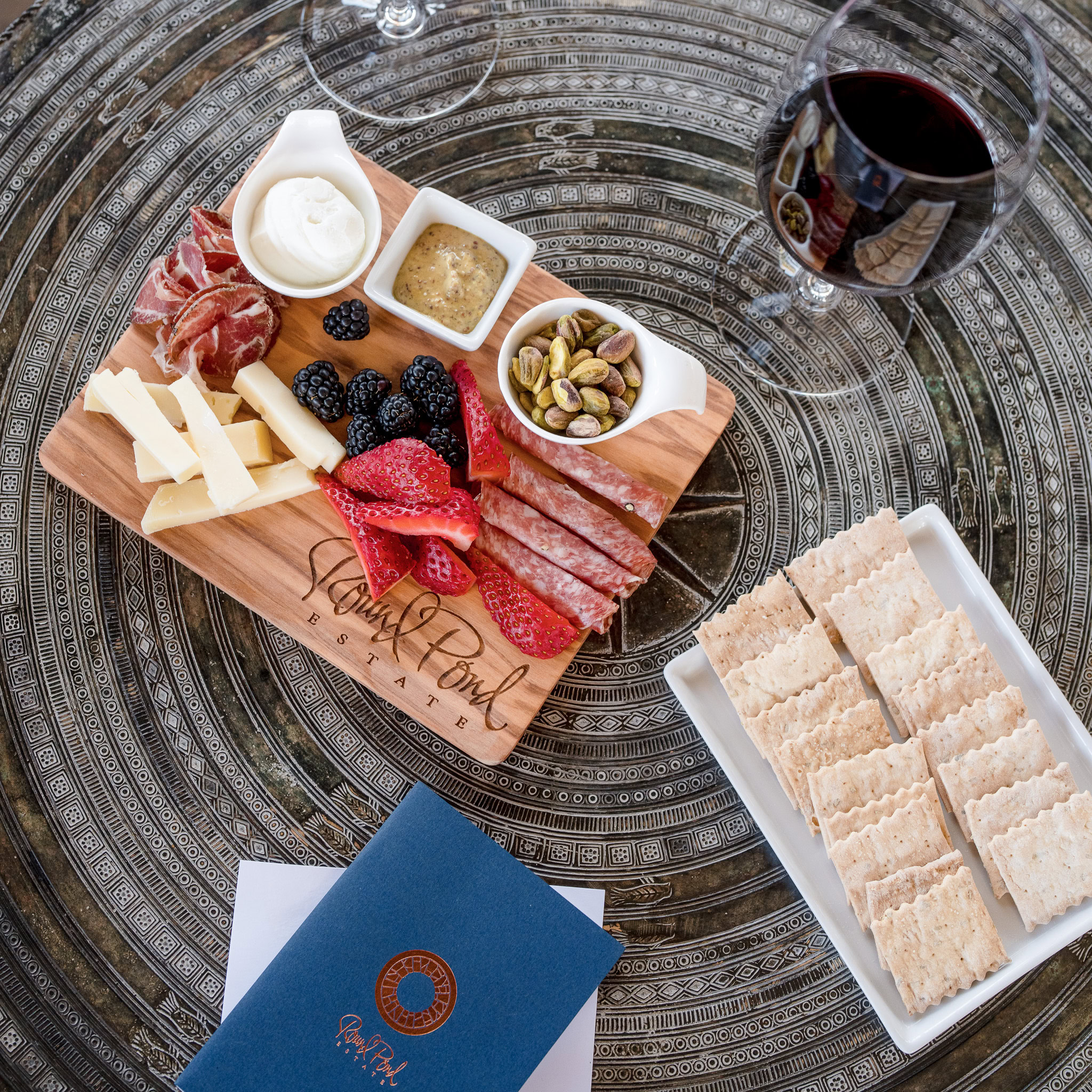 Charcuterie board, crackers, and a glass of red wine on grey table. Small blue notebook with the Round Pond Estate Logo in the bottom left corner.
