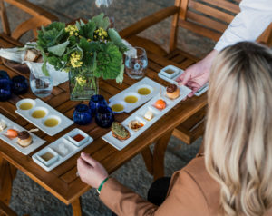 Blonde woman being served a white plate of small culinary bites with olive oil in small cups on a wood table.