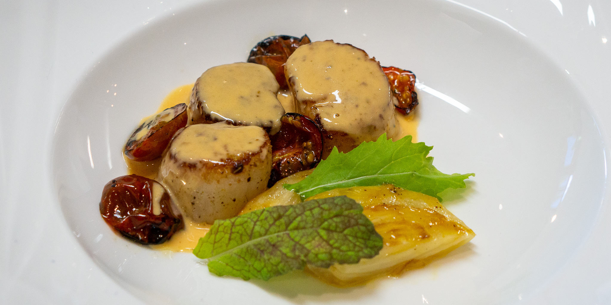 Summer Tomato Butter and Scallops White dish featuring three scallops covered in a tomato butter sauce granished with shiso leaf