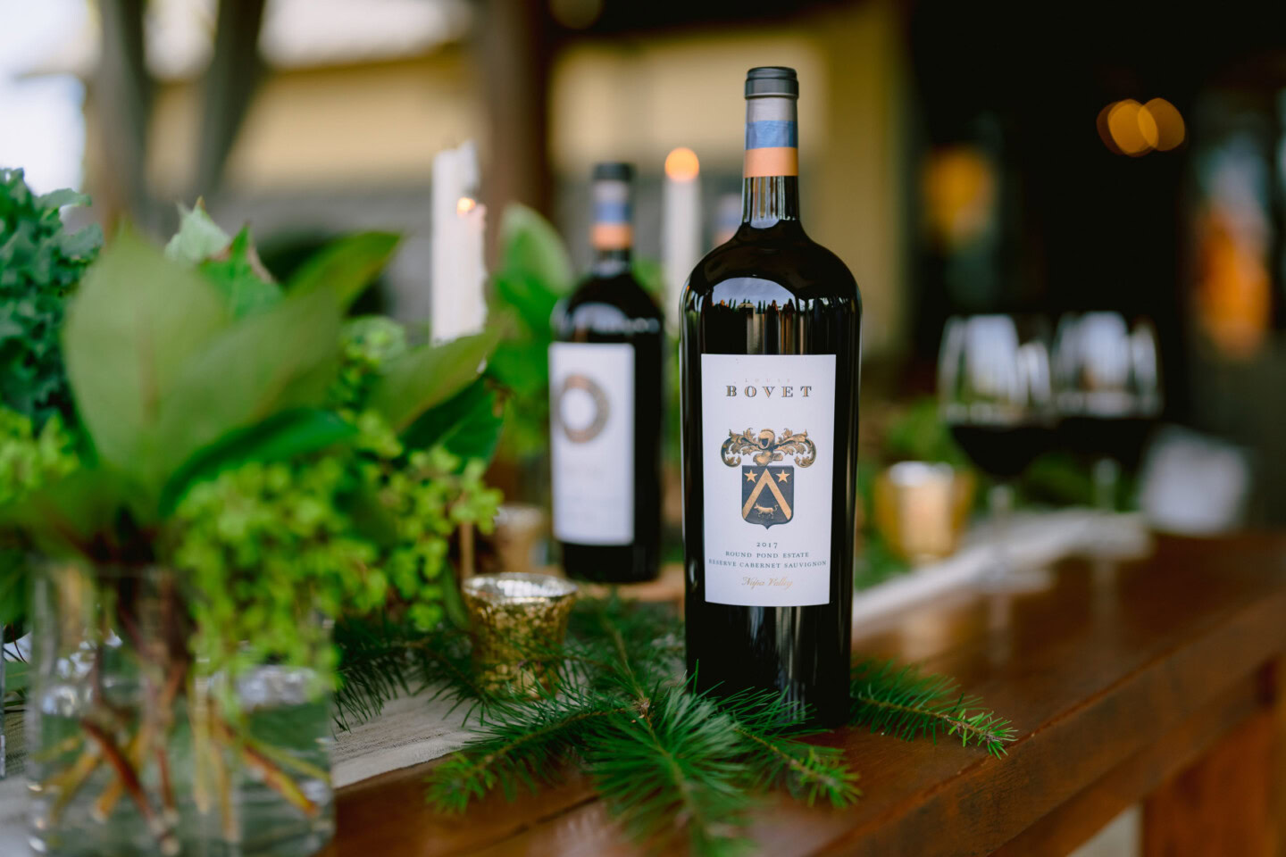 Elegant wine bottles on a wooden table at Round Pond Estate, surrounded by greenery and candles.