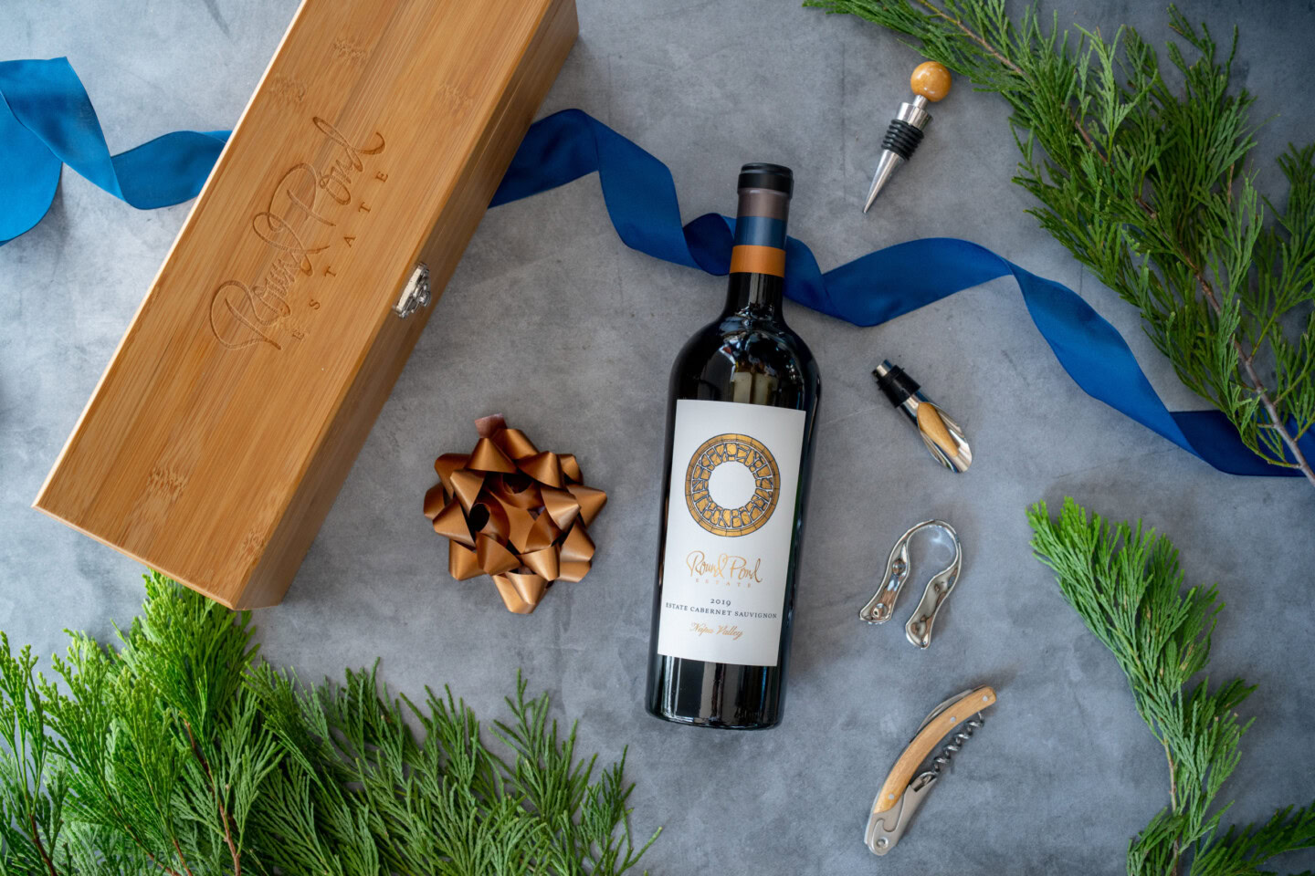 Wine bottle with a decorative label, surrounded by a wooden box, corkscrew, and festive greenery.