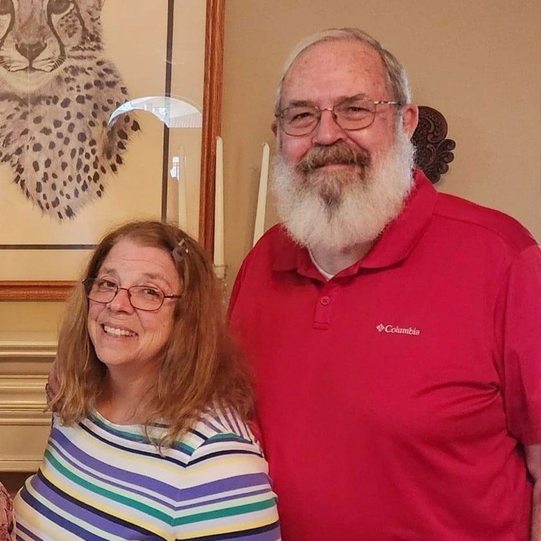 A smiling woman with glasses and a man with a beard, both in casual attire, standing indoors with a cheetah artwork in the back.