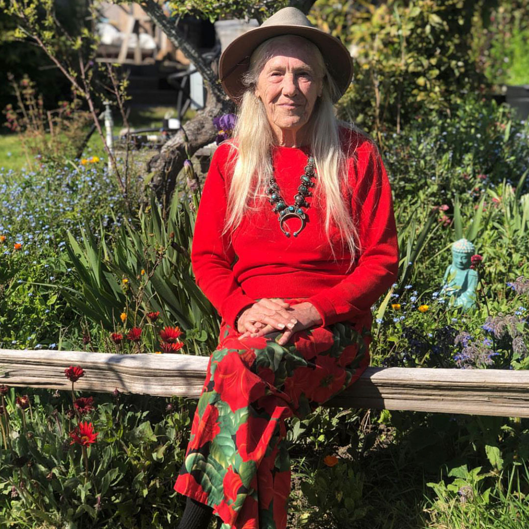 Elderly woman in a red dress and hat sitting in a lush garden with colorful flowers.