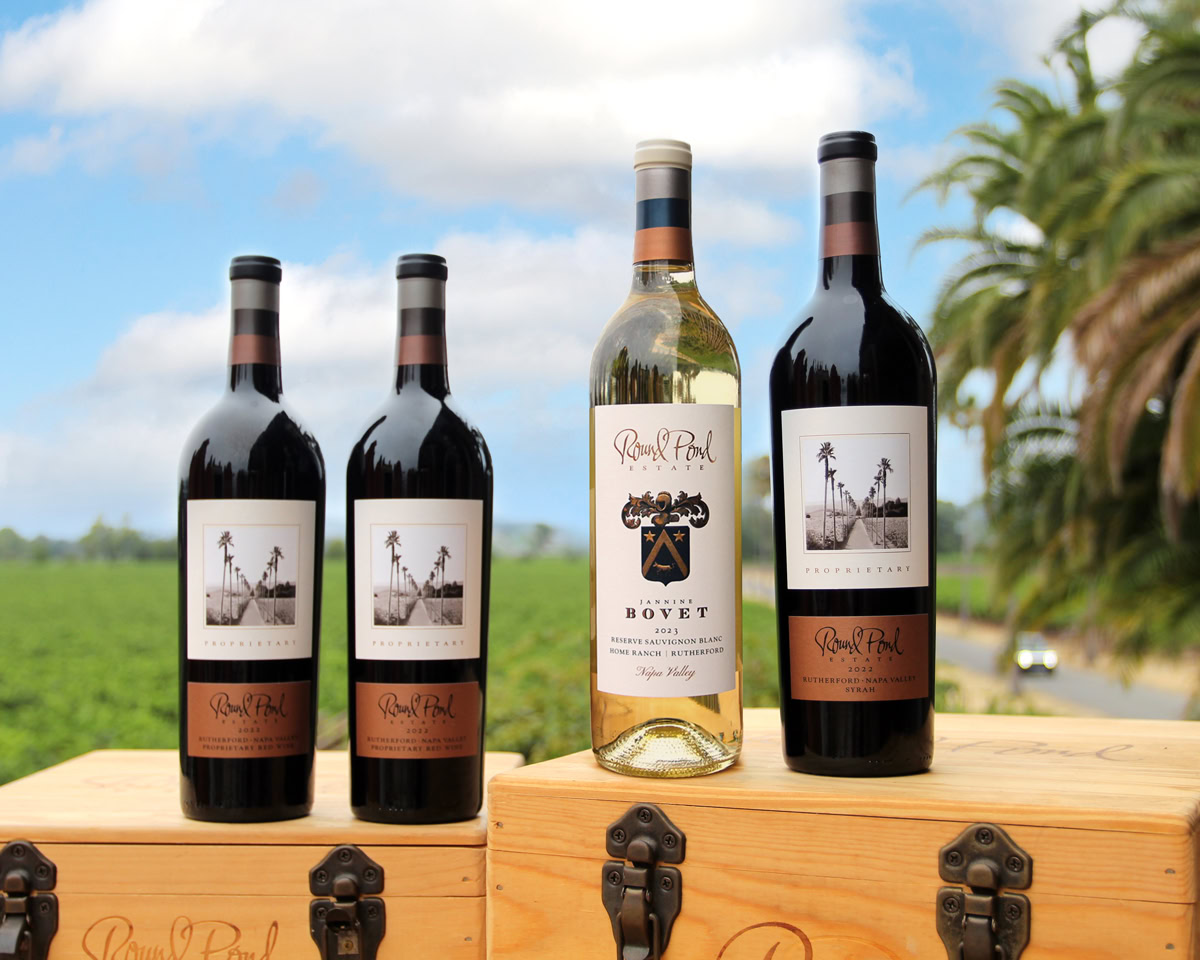Four bottles of wine from Round Pond Estate, two red and one white, displayed outdoors on a wooden box with a scenic backgrou.