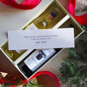 Gift note laid over a bottle of red and a bottle of white wine in a gift box with greenery and a red ribbon