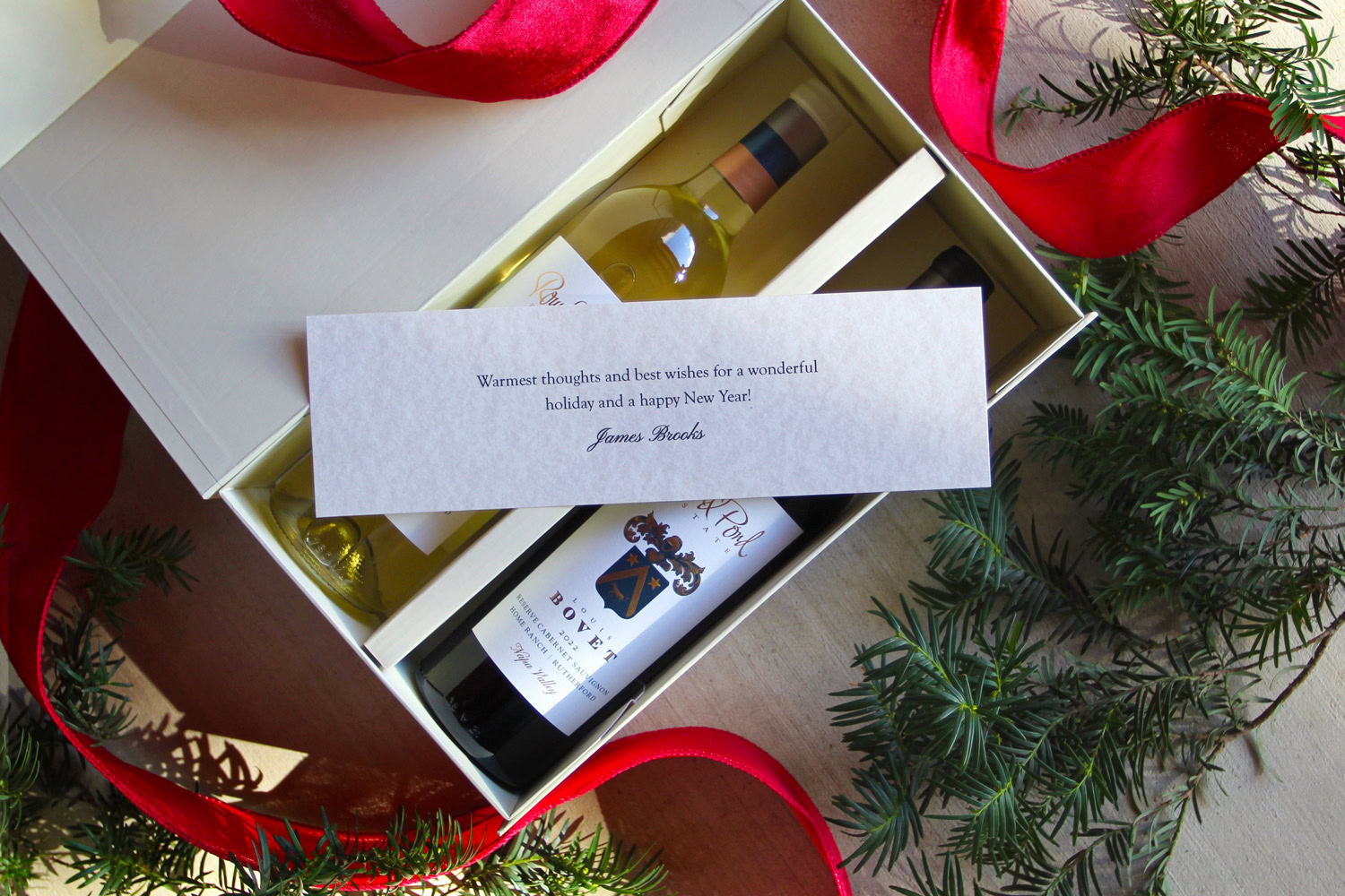A gift box containing a bottle of white wine, a bottle of red wine, and a holiday greeting card, surrounded by festive greene.