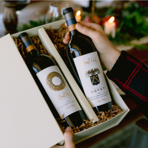 Hand removing a bottle of red wine from a gift box that includes another bottle of red wine with a candlelit table in the background