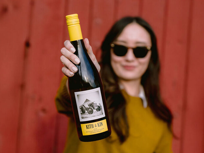 A woman wearing sunglasses and a mustard-colored sweater holds out a bottle of Kith & Kin 2023 Russian River Valley Chardonnay. The bottle has a yellow cap and a label featuring a black-and-white image of a tractor. She stands in front of a rustic red wooden wall, slightly out of focus, while the bottle is prominently in focus.