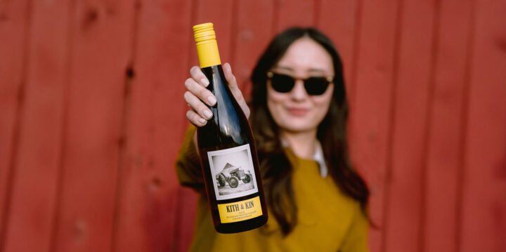 A woman wearing sunglasses and a mustard-colored sweater holds out a bottle of Kith & Kin 2023 Russian River Valley Chardonnay. The bottle has a yellow cap and a label featuring a black-and-white image of a tractor. She stands in front of a rustic red wooden wall, slightly out of focus, while the bottle is prominently in focus.