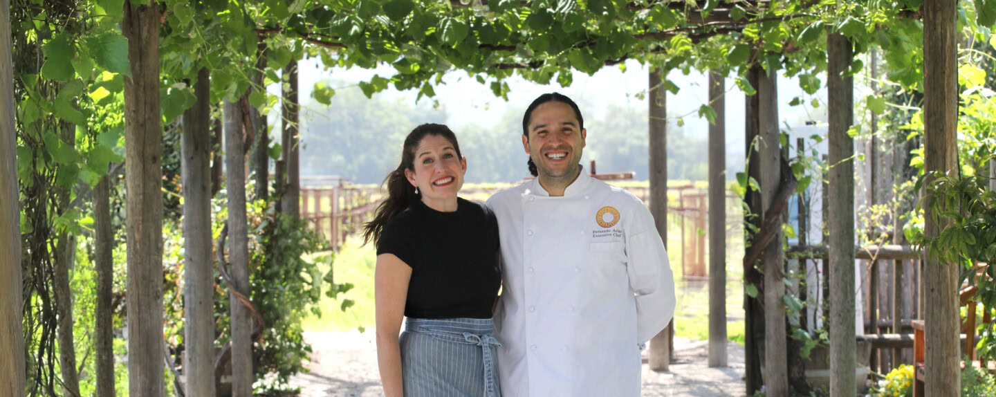 Smiling woman and chef standing under grapevine arbor at Round Pond Estate, showcasing vineyard hospitality and scenic outdoo.