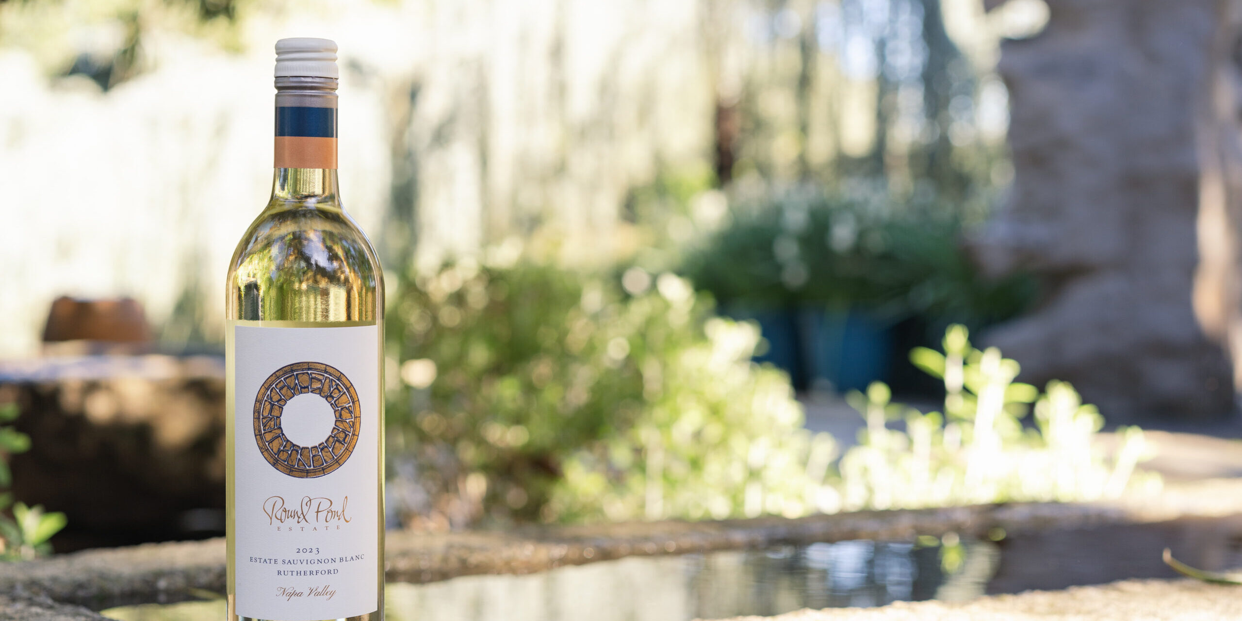 Bottle of Estate Sauvignon Blanc sitting on a stone edge of a fountain with blurry greenery behind it