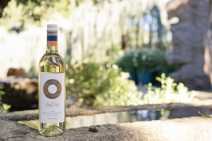 Bottle of Estate Sauvignon Blanc sitting on a stone edge of a fountain with blurry greenery behind it