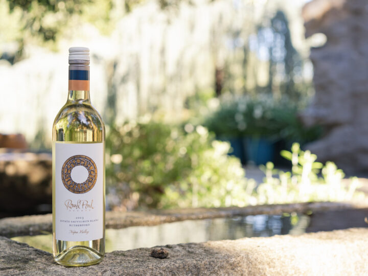 Bottle of Estate Sauvignon Blanc sitting on a stone edge of a fountain with blurry greenery behind it