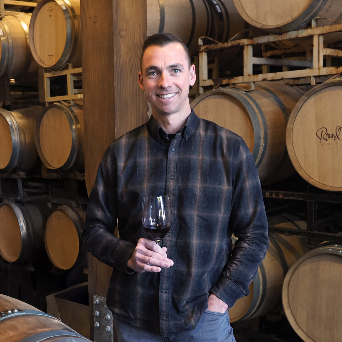 Jeff Plant, Estate Winemaker at Round Pond Estate, holding a glass of red wine in the barrel room. Surrounded by stacks of French oak barrels, Jeff crafts wines that reflect the character of Napa Valley with precision and care.