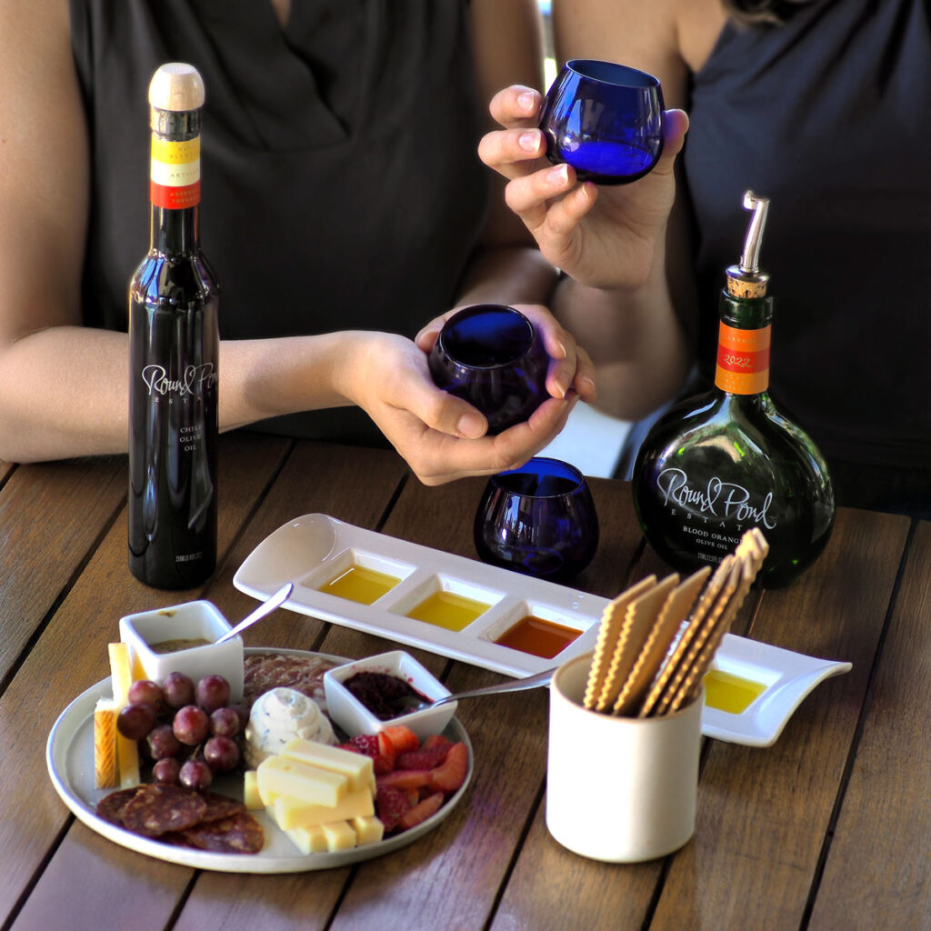 Guests enjoying Round Pond Estate's Vino & Olio Experience in Napa Valley, featuring a premium olive oil tasting with signature blue glasses, a curated wine tasting, and an artisan charcuterie board with cheese, cured meats, fresh fruit, and gourmet spreads — a must-try Napa Valley culinary experience.