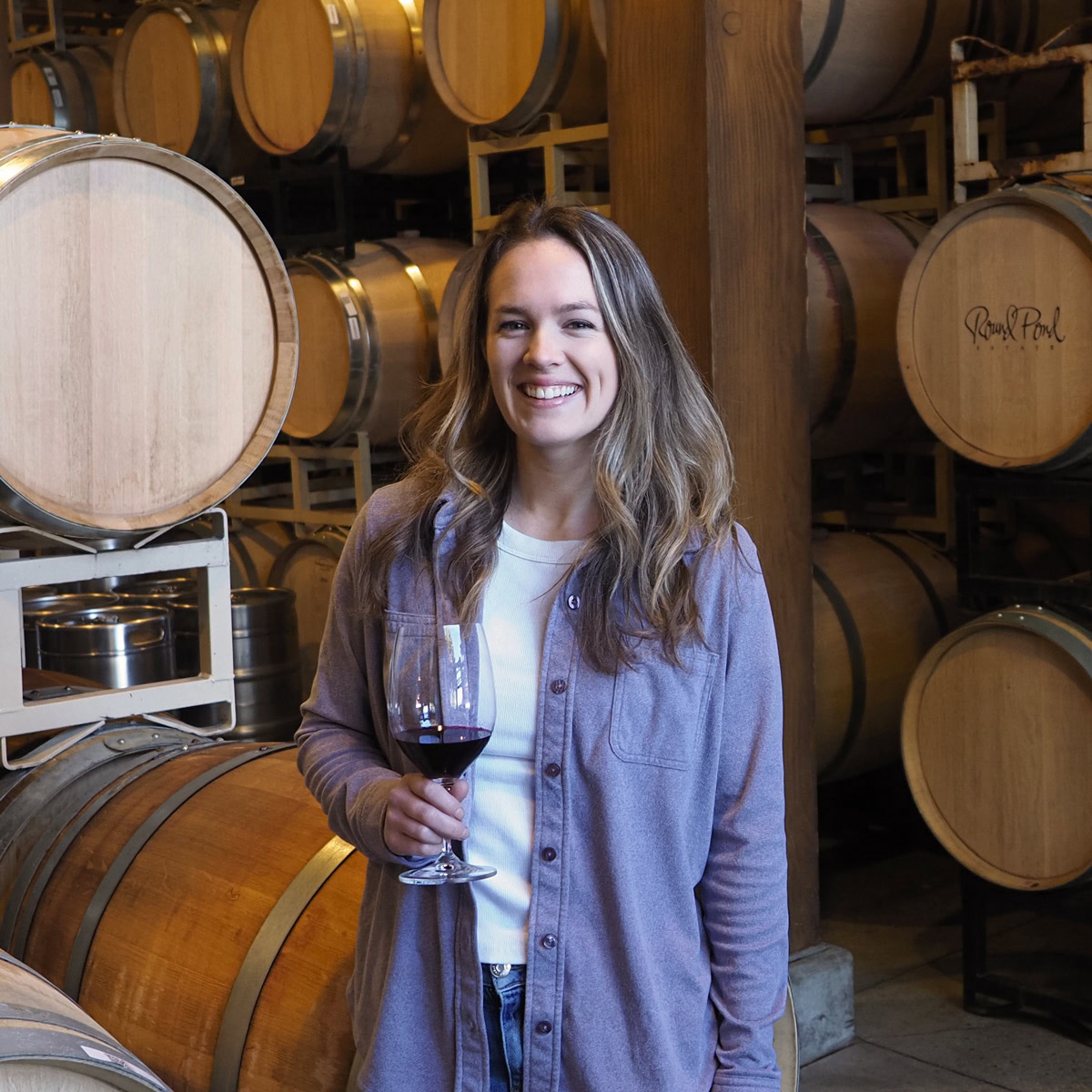Rachel Jess, Assistant Winemaker at Round Pond Estate, smiling with a glass of red wine in hand in the estate barrel room. Surrounded by stacks of aging oak barrels, Rachel brings thoughtful craftsmanship and energy to Napa Valley winemaking.