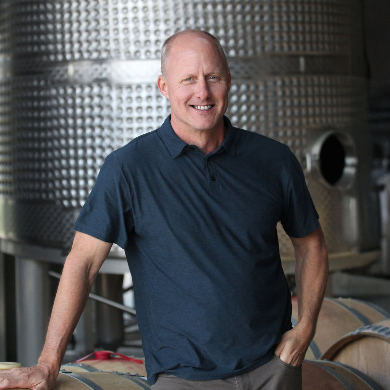 Thomas Rivers Brown, consulting winemaker at Round Pond Estate, standing in the winery cellar with stainless steel fermentation tanks and wine barrels in the background. Napa Valley winemaking expert known for crafting award-winning wines.