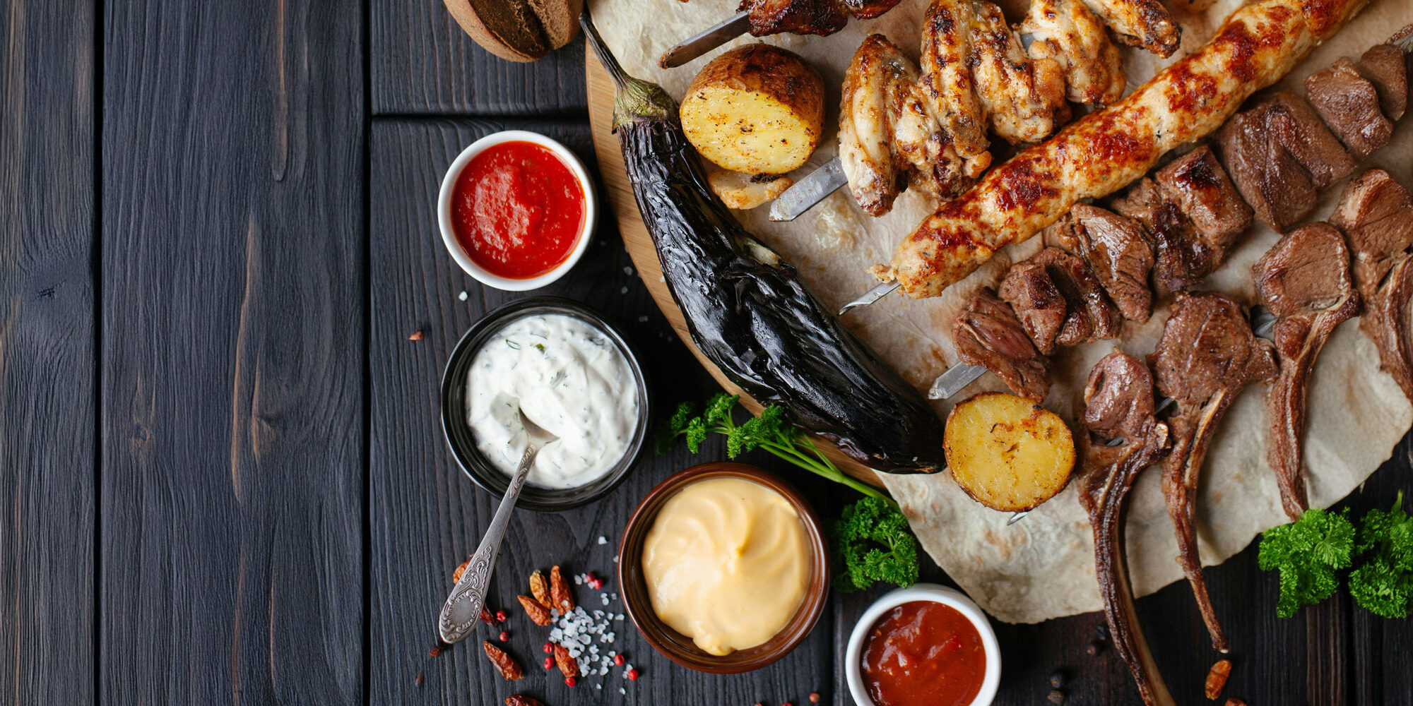 Chive Aioli Recipe A rustic wooden table topped with a grilled feast including lamb chops, chicken wings, skewered meats, grilled eggplant, and potatoes, surrounded by bowls of colorful sauces—featuring a creamy chive aioli at the center, served as a dip for grilled meats.