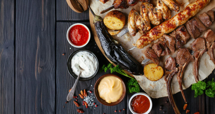 Chive Aioli Recipe A rustic wooden table topped with a grilled feast including lamb chops, chicken wings, skewered meats, grilled eggplant, and potatoes, surrounded by bowls of colorful sauces—featuring a creamy chive aioli at the center, served as a dip for grilled meats.