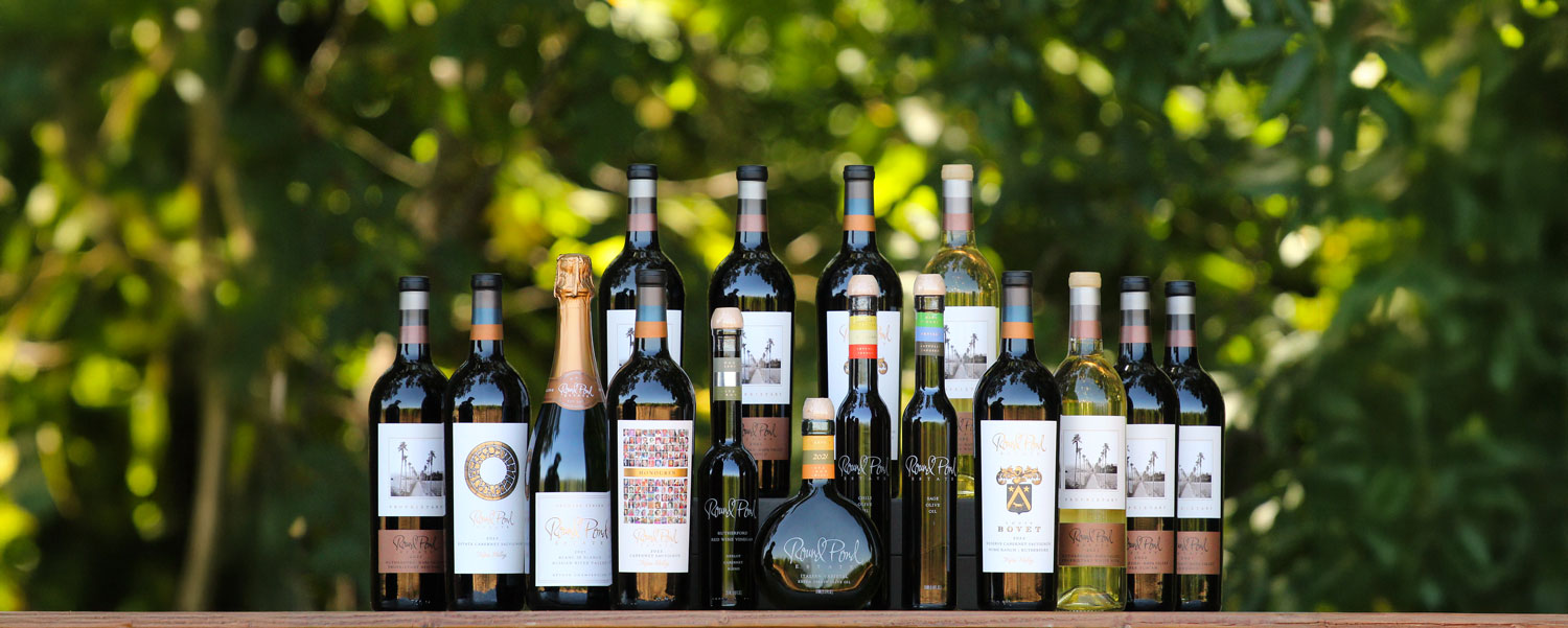 Round Pond Estate Wines A collection of Round Pond Estate wines and olive oils arranged on a wooden table outdoors with a background of lush green trees in soft focus.
