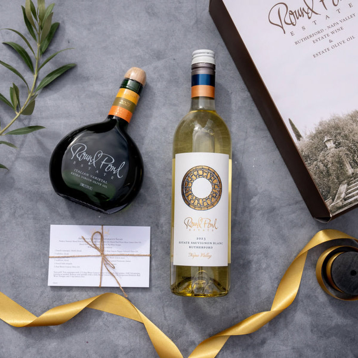 Two bottles of wine and olive oil from Round Pond Estate on a gray surface with a box and ribbon.