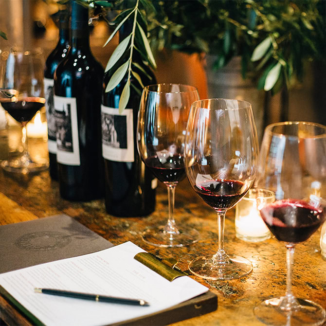 Three glasses of red wine on a wooden table with wine bottles and greenery in the background.