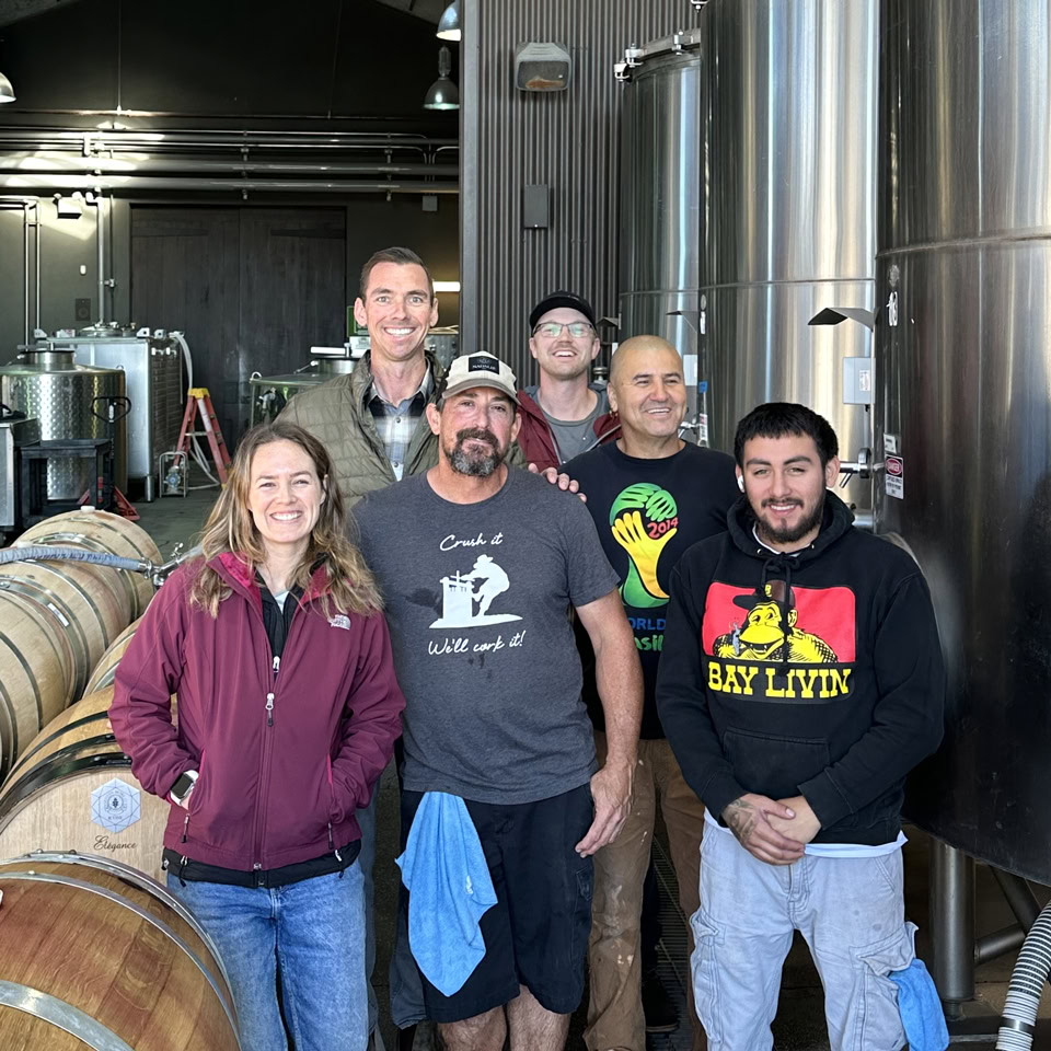 Group of six people at Round Pond Estate winery, smiling inside the production area with barrels and stainless steel tanks.