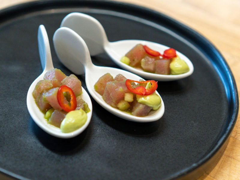 Close-up of Tuna Aguachile served on white spoons with chili and avocado, presented on a black tray.