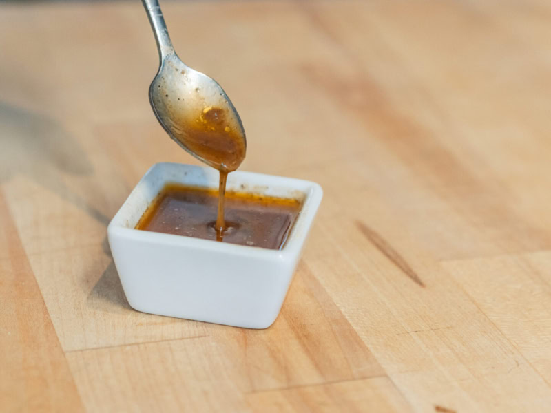 A spoon drizzling tomato dressing into a small white square dish on a wooden surface.