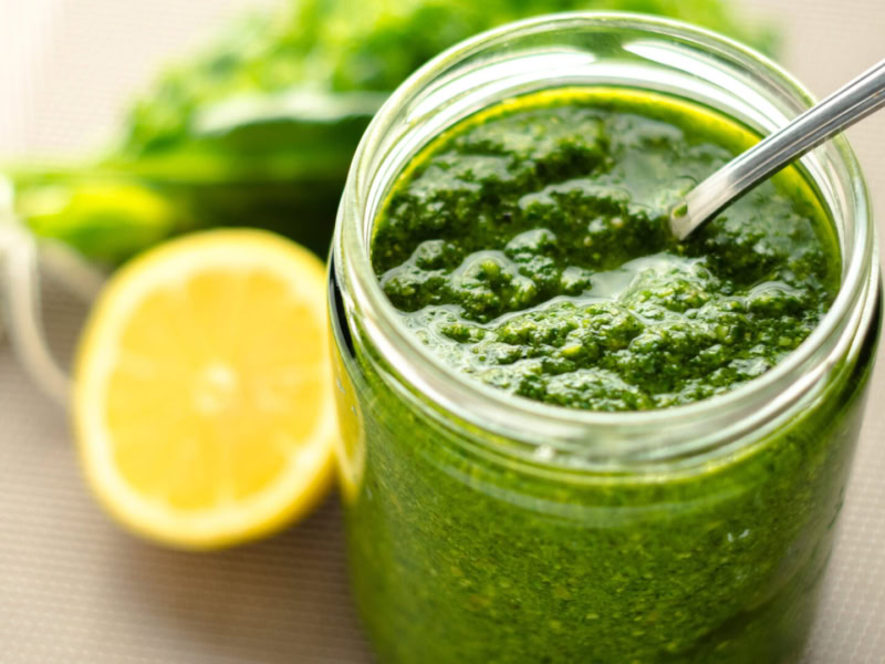 Fresh green harissa sauce in a glass jar with a spoon, lemon half, and herbs in the background.