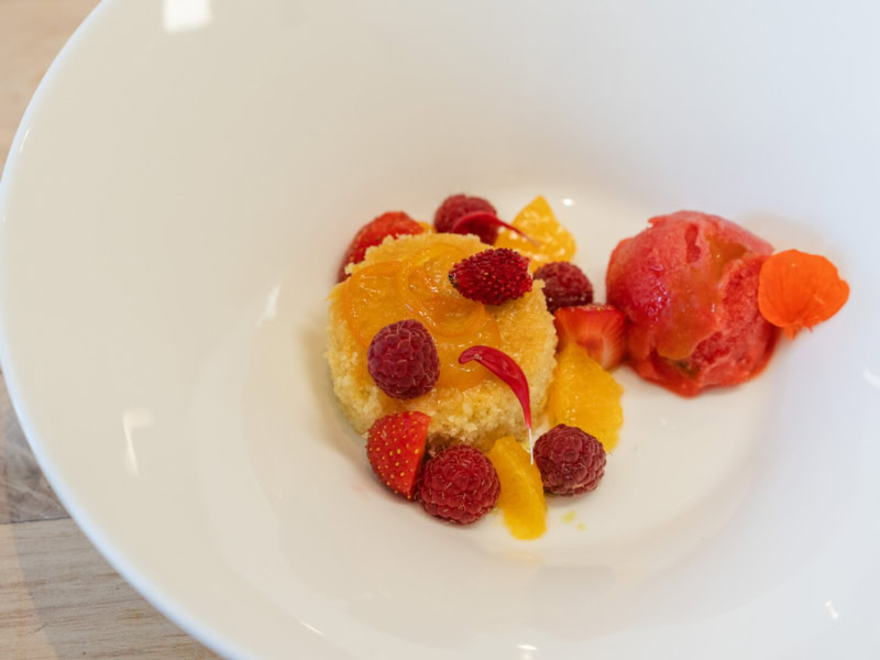 Meyer Lemon Cake topped with raspberries, strawberries, and orange slices, served with a fruit sorbet on a white plate.