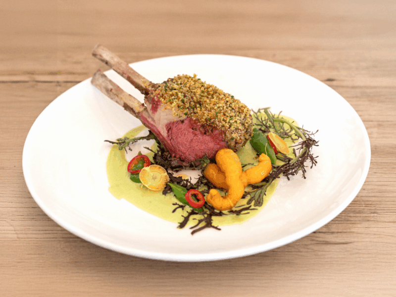 Herbed lamb chop with a crust of herbs, served with peas, carrots, and a green sauce on a white plate.