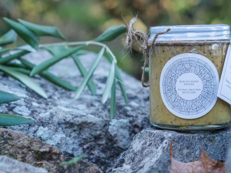 Stone Ground Mustard in a glass jar with a rustic label, placed on a rock outdoors with greenery in the background.