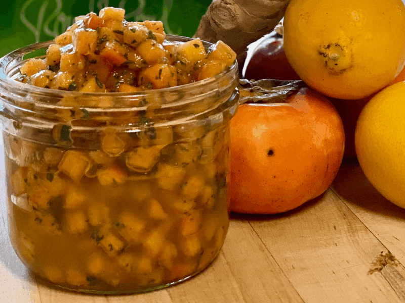 Close-up of a jar filled with bright orange persimmon relish, surrounded by fresh apples on a wooden surface.